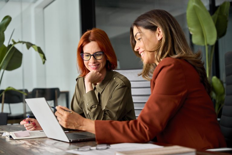 Two women sit at a desk in an office, smiling at a laptop screen. One gestures as they discuss, reflecting a positive work culture. Green plants add warmth to the background.