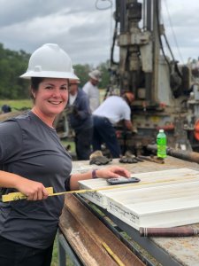 A woman wearing a white hard hat and gray shirt smiles while measuring core samples with a tape measure at an Orlando outdoor drilling site. Workers and drilling equipment are visible in the background.