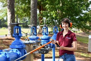 A woman wearing orange earmuffs and a maroon shirt stands outdoors by large blue industrial valves in Orlando, holding a measuring instrument and smiling at the camera. Trees and greenery are in the background.