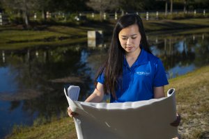 A woman in a blue shirt stands outdoors by a pond in Orlando, examining a large rolled-out paper, possibly a blueprint or map, with trees and a grassy area in the background.
