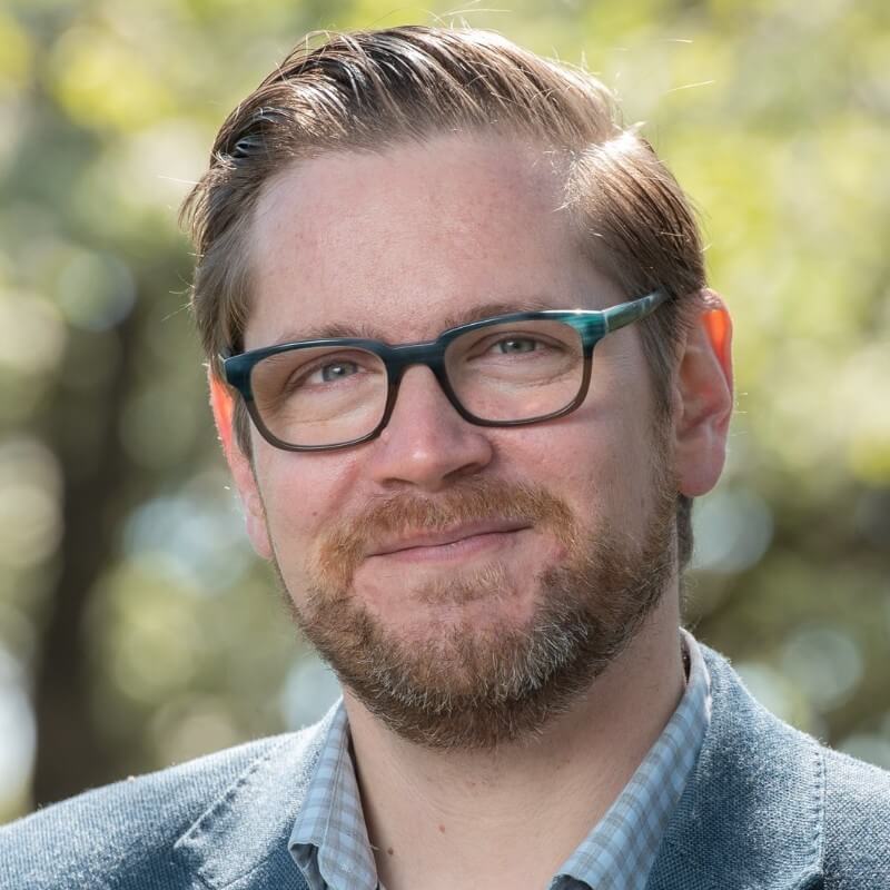 A man with light brown hair and a beard, wearing glasses, a light-colored shirt, and a gray blazer, smiles outdoors with blurred green foliage in the background. A man with light brown hair and a beard, wearing glasses, a light-colored shirt, and a gray blazer, smiles outdoors with blurred green foliage in the background.