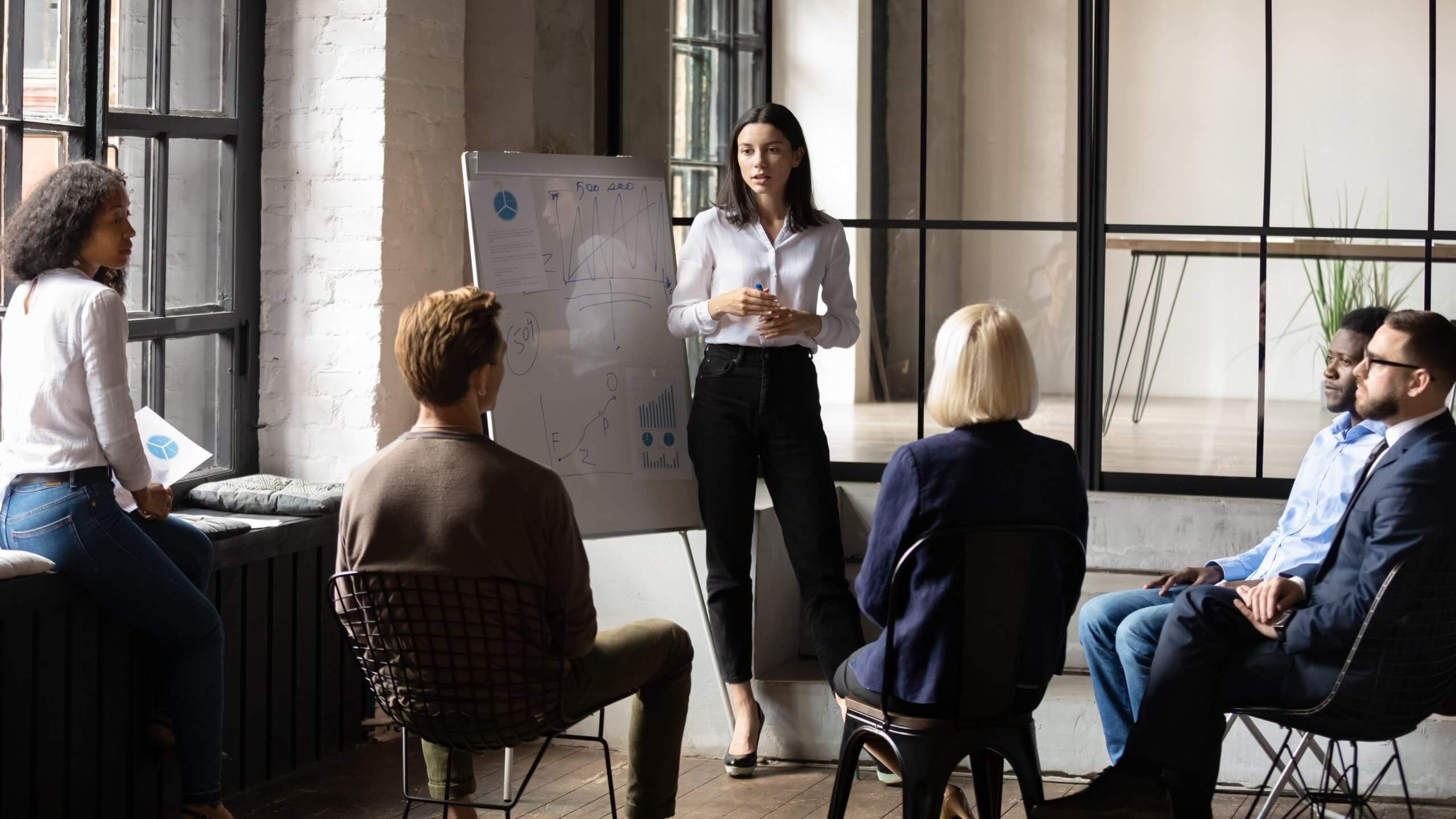 A woman stands by a whiteboard giving a presentation on public sector strategies to five seated colleagues in a modern office with large windows and industrial-style decor. A woman stands by a whiteboard giving a presentation on public sector strategies to five seated colleagues in a modern office with large windows and industrial-style decor.