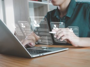 A person using a laptop at a desk interacts with virtual screens displaying digital profiles and checklists, representing data analysis or career recruitment.