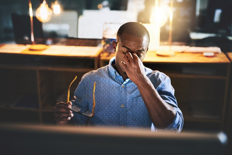 A man sits at a desk holding his glasses and pinching the bridge of his nose, appearing tired or stressed, with a computer screen displaying performance data and warm lighting in the background.