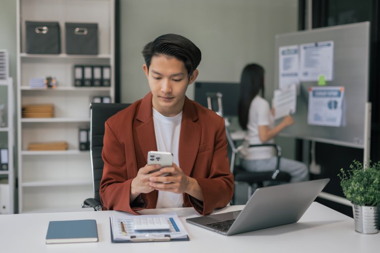 A man in a red blazer sits at a desk using his smartphone, with a laptop, notebook, and clipboard in front of him. In the background, a woman works at a whiteboard in a modern public sector office setting.