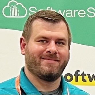 A man with short brown hair and a beard smiles at the camera. He is wearing a teal collared shirt and a lanyard. A green and white banner with the word "Software" is visible in the background. A man with short brown hair and a beard smiles at the camera. He is wearing a teal collared shirt and a lanyard. A green and white banner with the word "Software" is visible in the background.