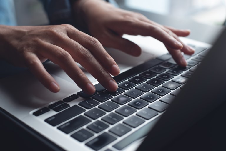 Close-up of hands typing on a laptop keyboard, with natural light illuminating the scene from the side. The person appears to be working or advancing their career on the computer.