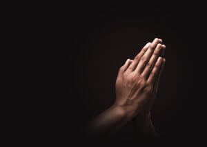 Two hands are pressed together in a prayer gesture against a dark background, with light highlighting the hands and creating a sense of focus, reverence, and the quiet determination that leads to success.