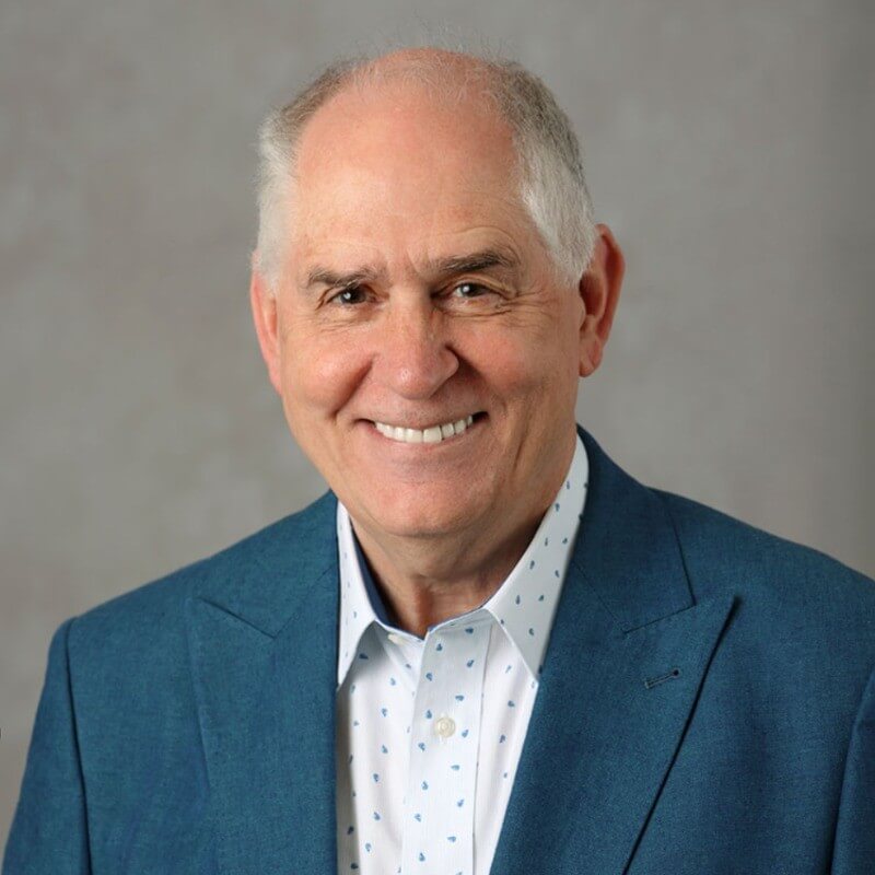 Smiling older man with short gray hair, wearing a teal blazer and a white shirt with a subtle pattern, posing in front of a plain, light gray background.