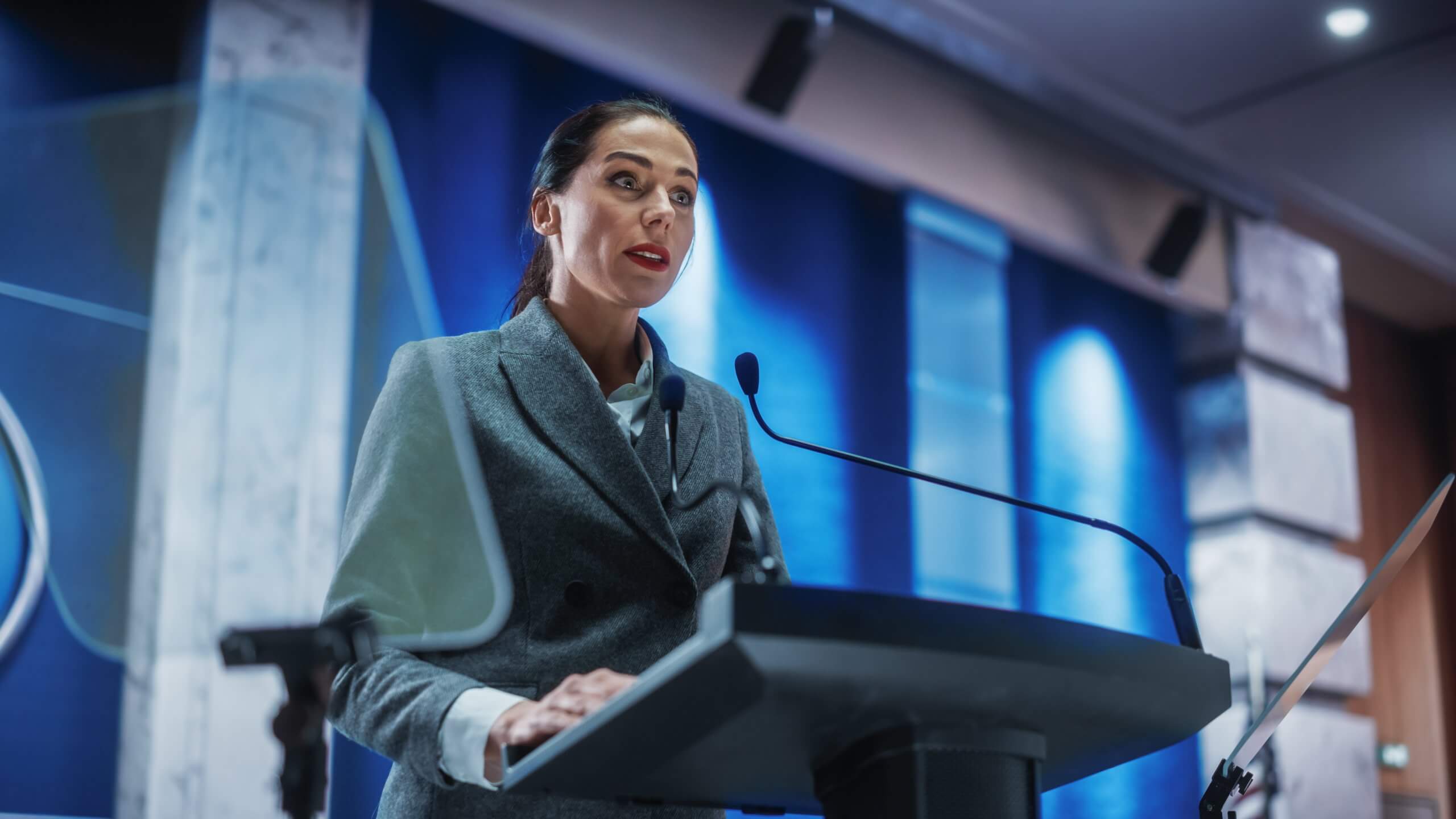 A woman in a gray blazer stands at a podium, demonstrating leadership as she speaks into microphones in a modern conference room with blue lighting and glass panels in the background.