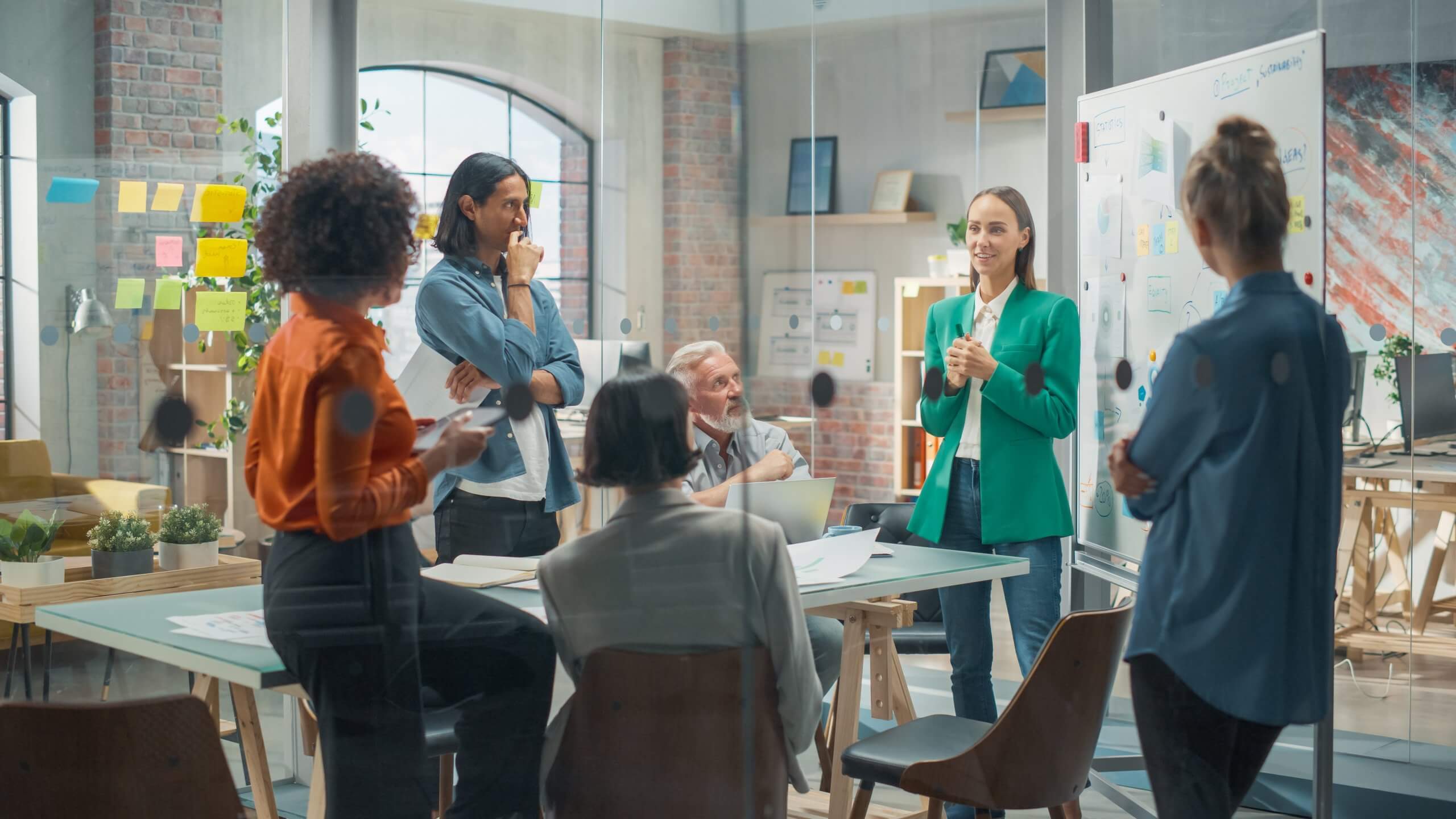 A group of five workforce professionals are having a meeting in a modern office. One person stands and presents at a whiteboard while the others sit or stand, listening and engaging in discussion. The room is bright and filled with natural light.