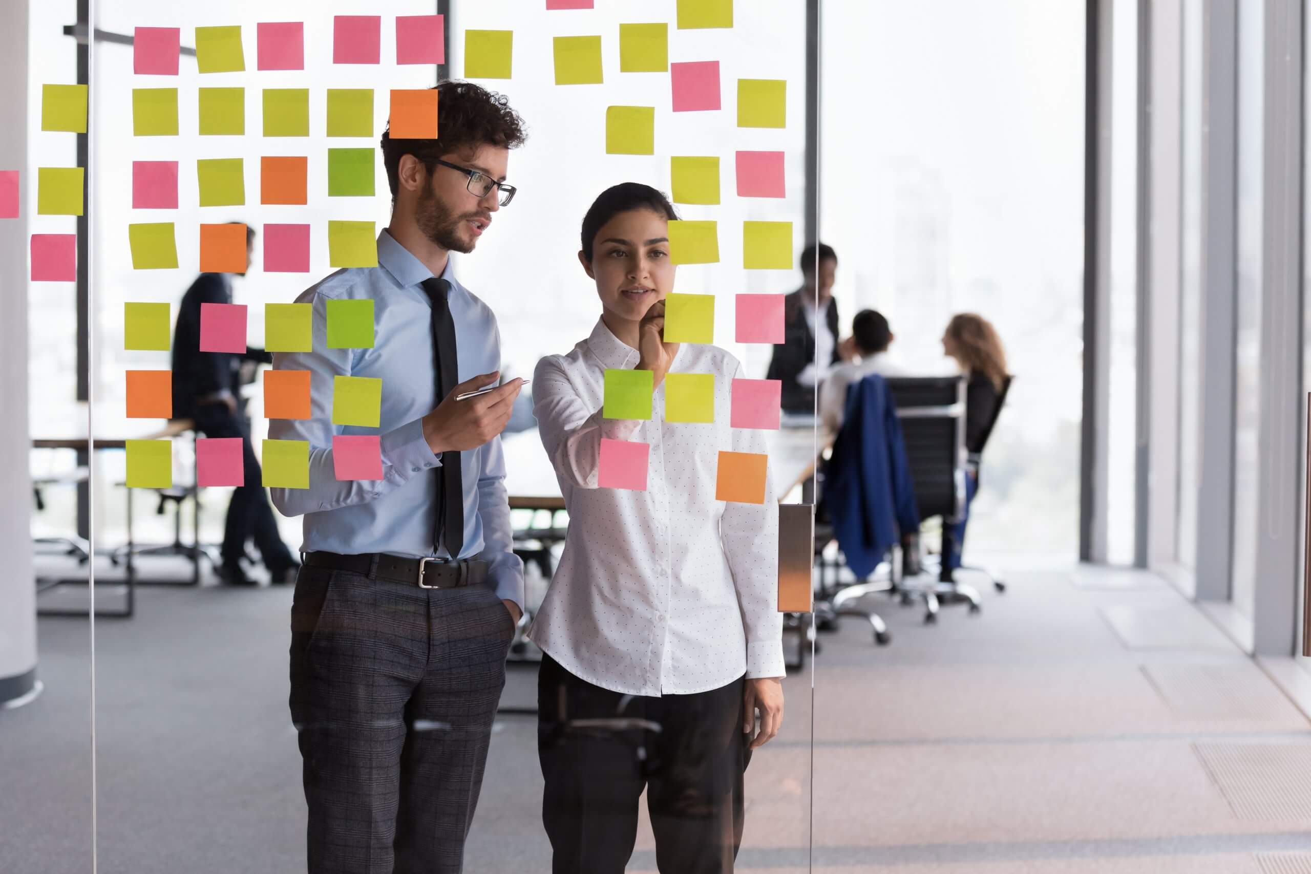 Two professionals stand by a glass wall covered with colorful sticky notes, discussing ideas in a modern office that reflects a collaborative culture. Other people are working at desks in the background.