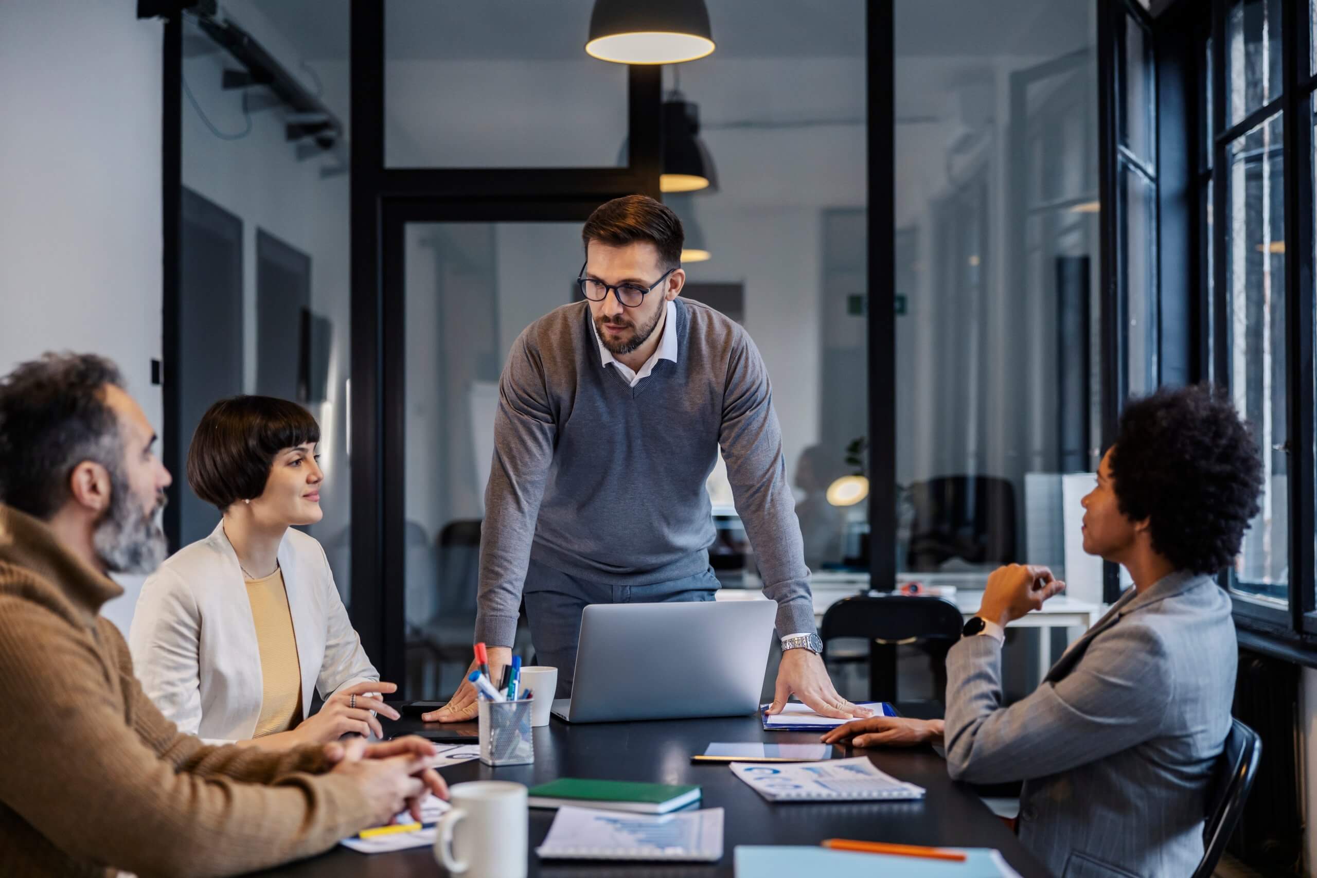 A woman stands by a whiteboard giving a presentation on public sector strategies to five seated colleagues in a modern office with large windows and industrial-style decor.