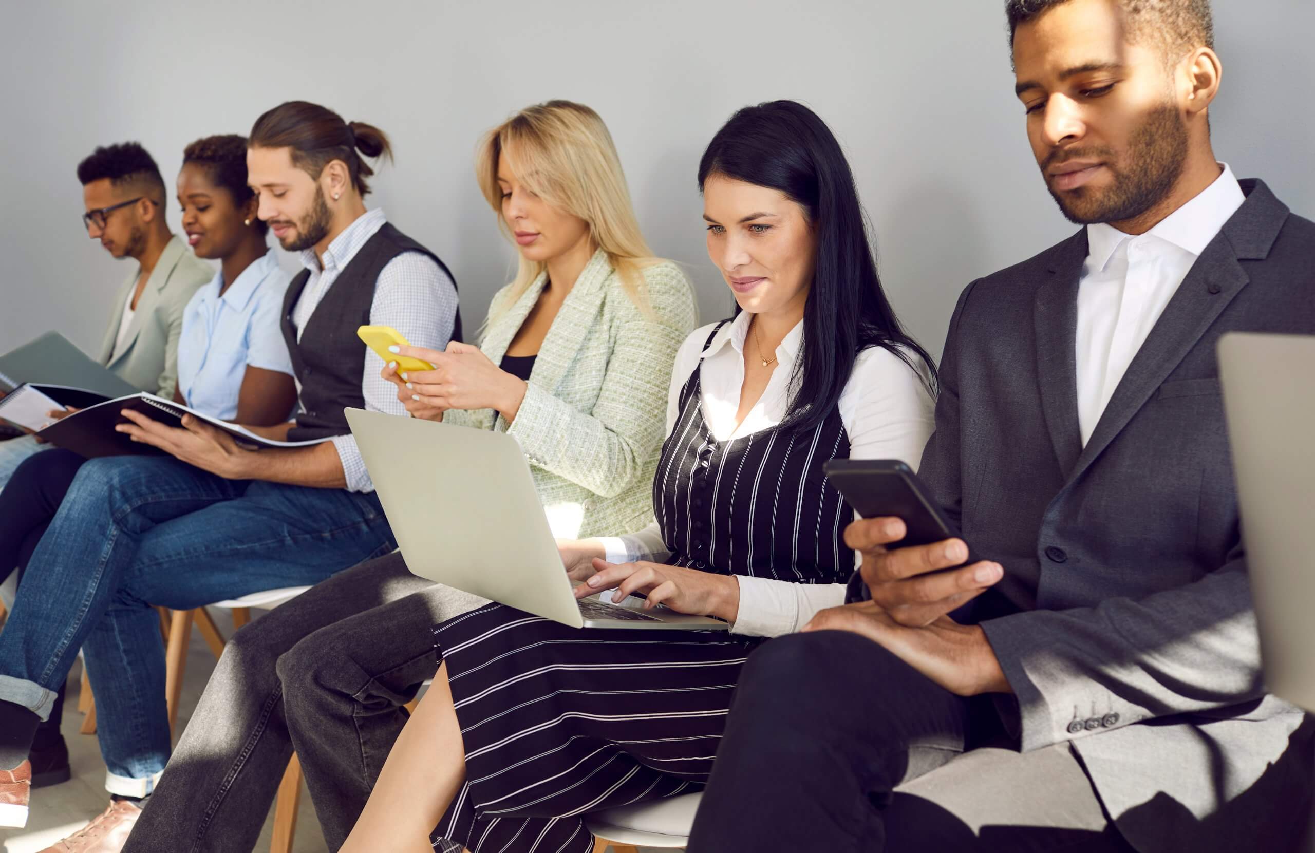 A diverse group of six professionals sits in a row, focusing on laptops, notebooks, and smartphones, possibly waiting for interviews or a public service business meeting. They appear engaged and dressed in business attire.