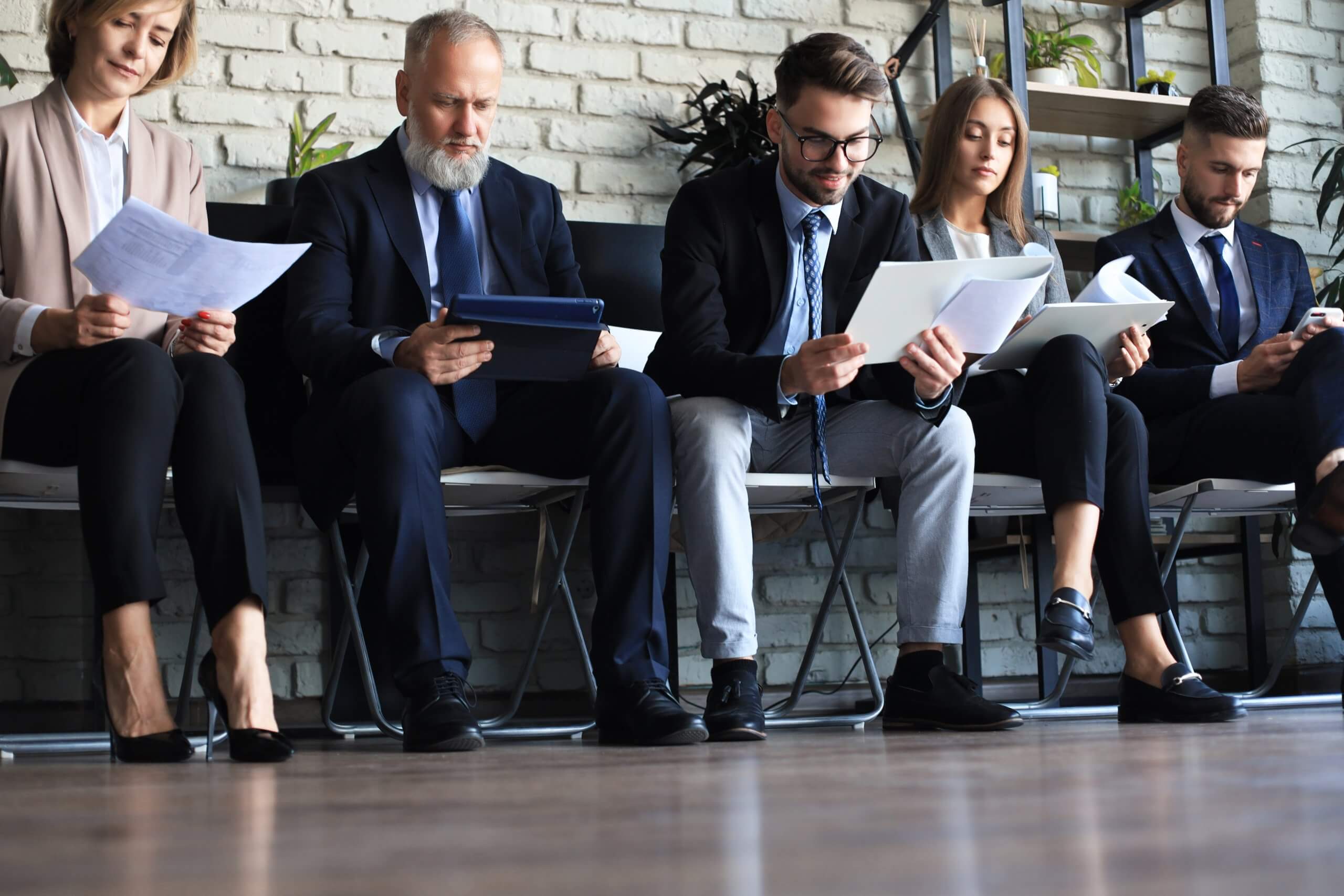Five professionally dressed people sit in a row on chairs, reviewing documents or using a tablet, possibly waiting for a job interview, engaged in quiet conversations against a light brick wall with plants in the background.