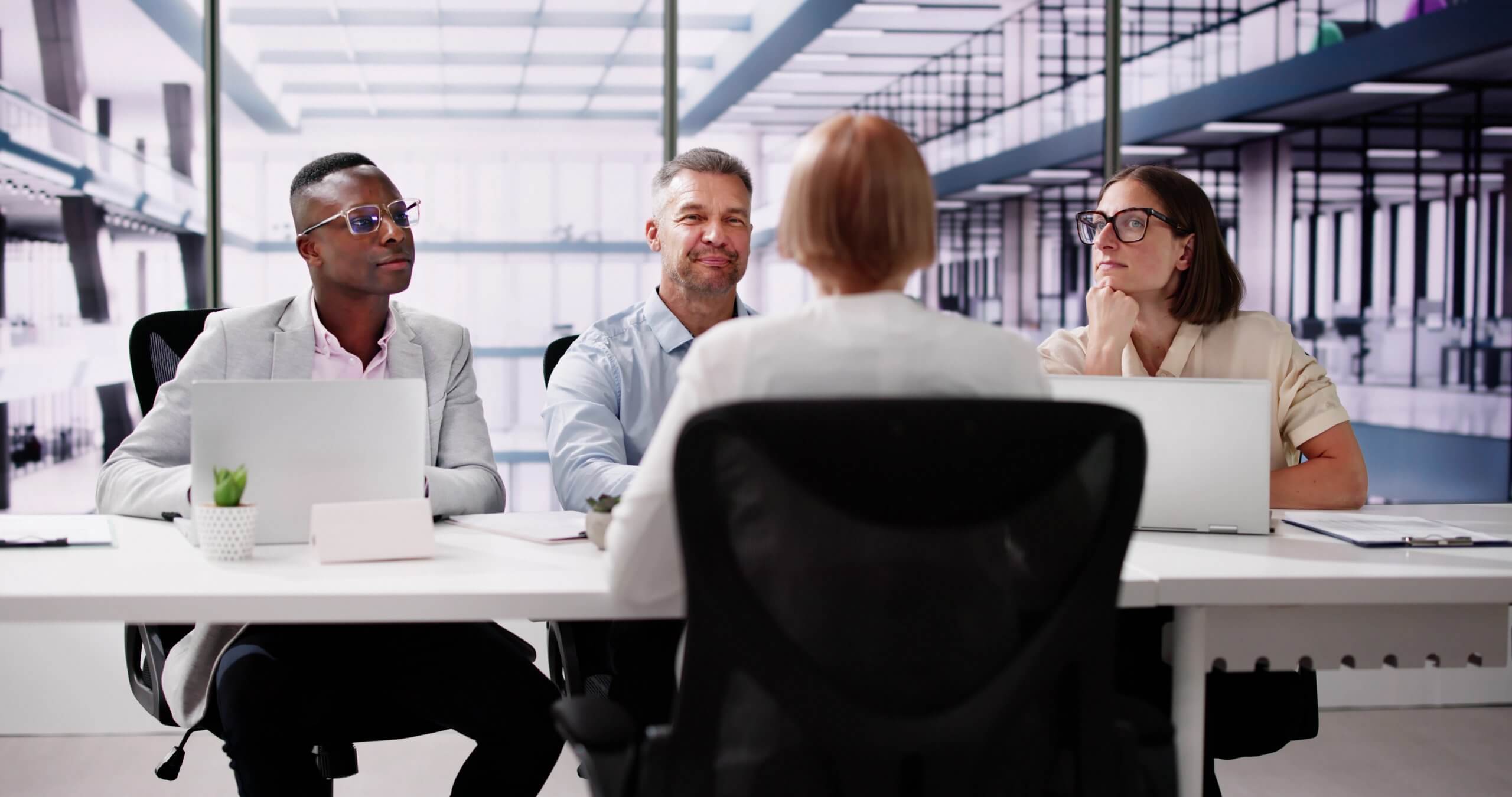 Four professionals sit at a conference table in a modern office, facing a woman with her back to the camera. Laptops and papers are on the table as they discuss leadership during an engaging meeting or interview.