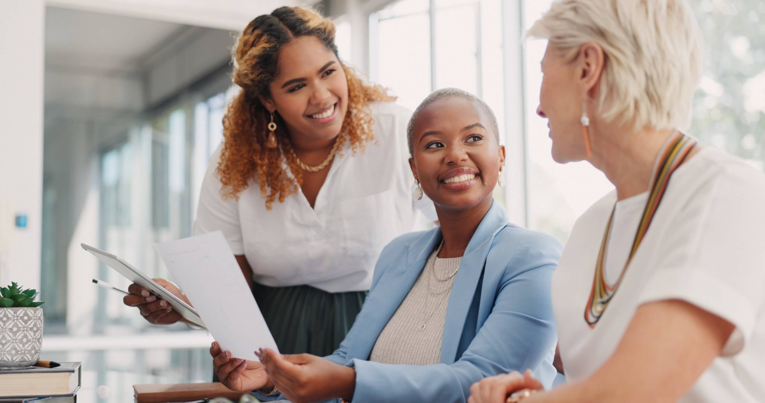 Three women in business attire sit together in a bright office, smiling and talking. One woman holds documents, while another leans over to join the conversation—a positive snapshot of a diverse workforce. A small plant sits on the table.