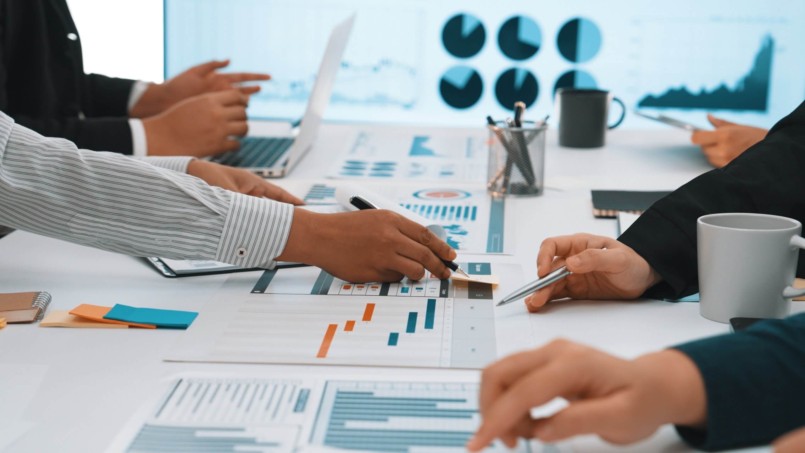 A group of people in business attire discuss charts and graphs spread on a conference table, using pens, papers, and laptops, reflecting a culture of collaboration with data visualizations displayed on a screen in the background.