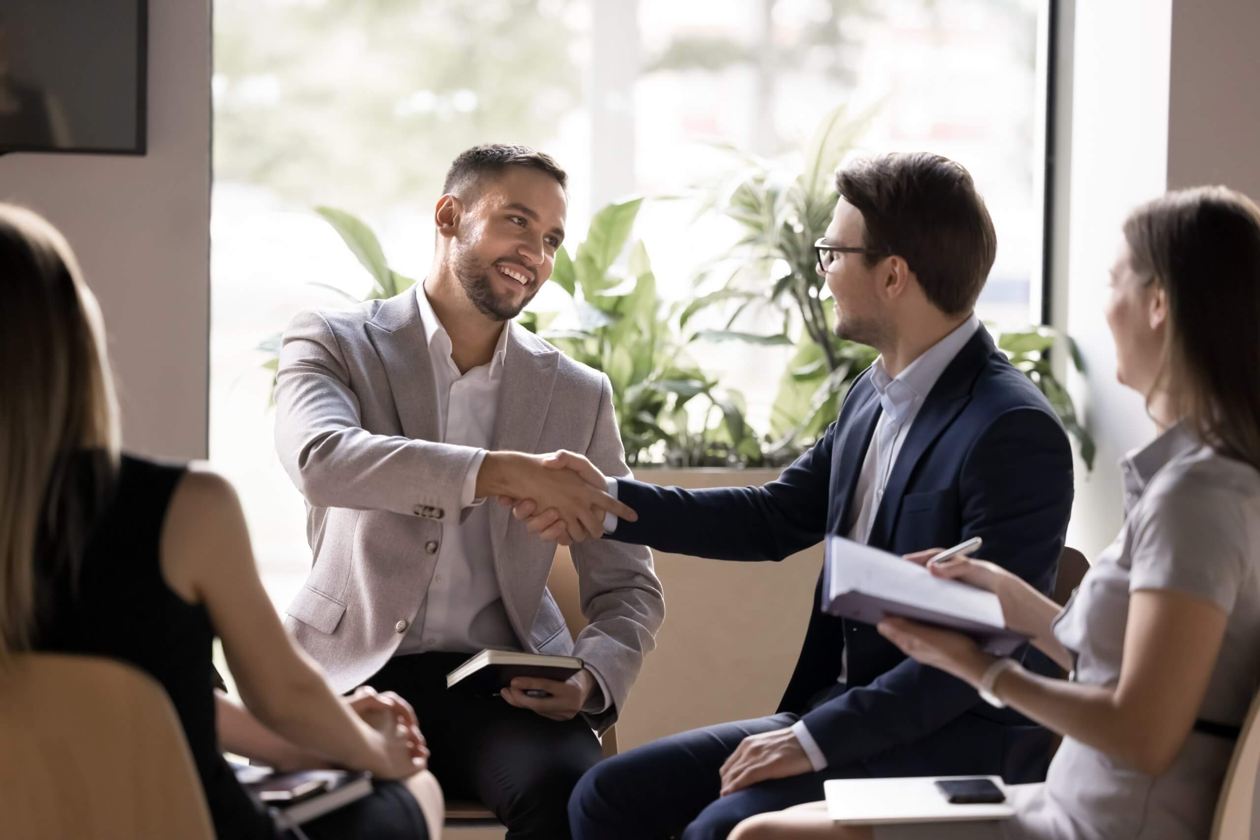 A woman in an orange blouse stands by a whiteboard covered with colorful notes and diagrams, smiling and presenting insights to two colleagues in a modern public sector office setting.
