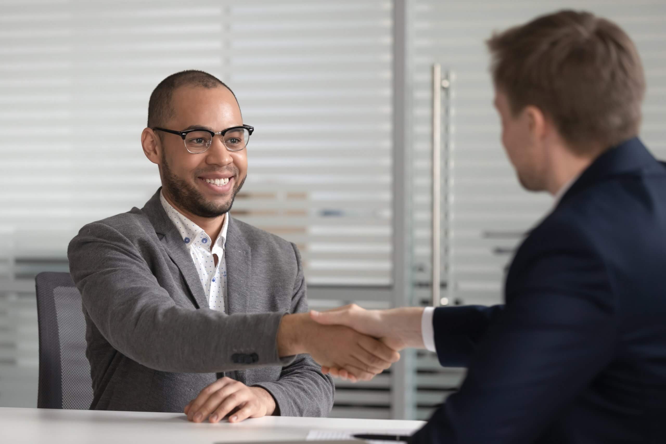 Two men in business attire sit at a table in an office, smiling and shaking hands, suggesting a successful public service meeting or agreement.