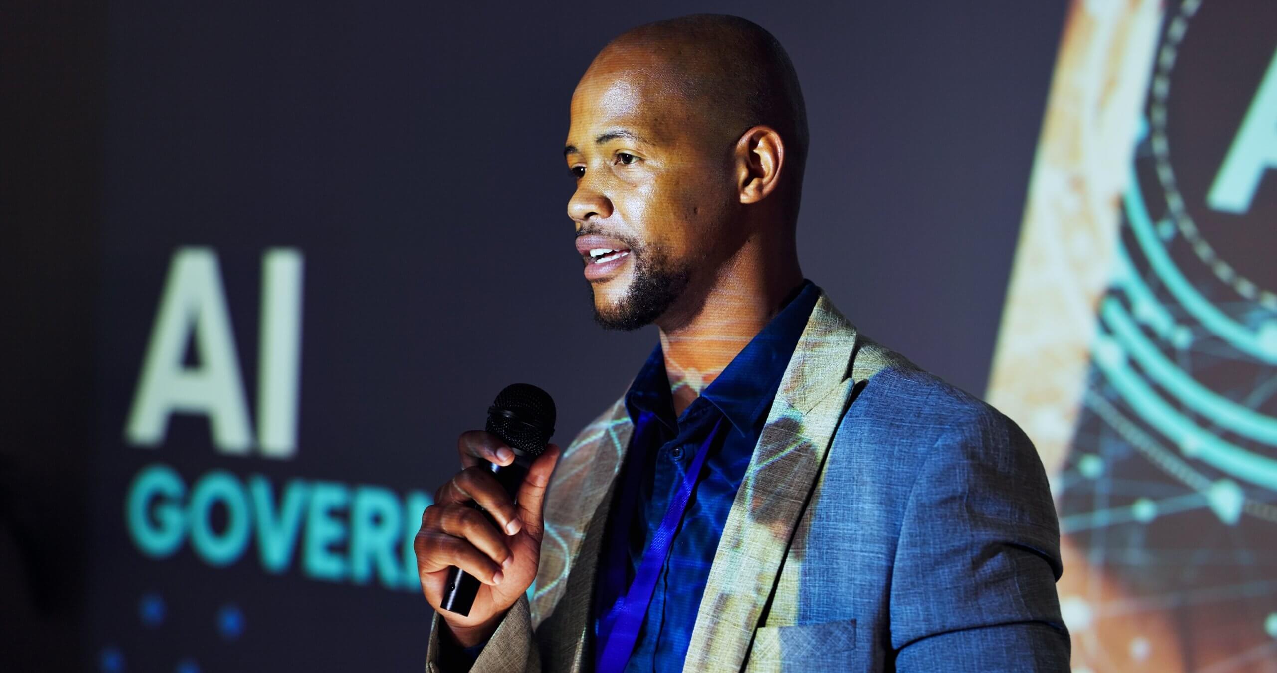 A man in a blazer holds a microphone and speaks on stage in front of a screen displaying the words "AI GOVERN," addressing key topics about AI in the public sector.