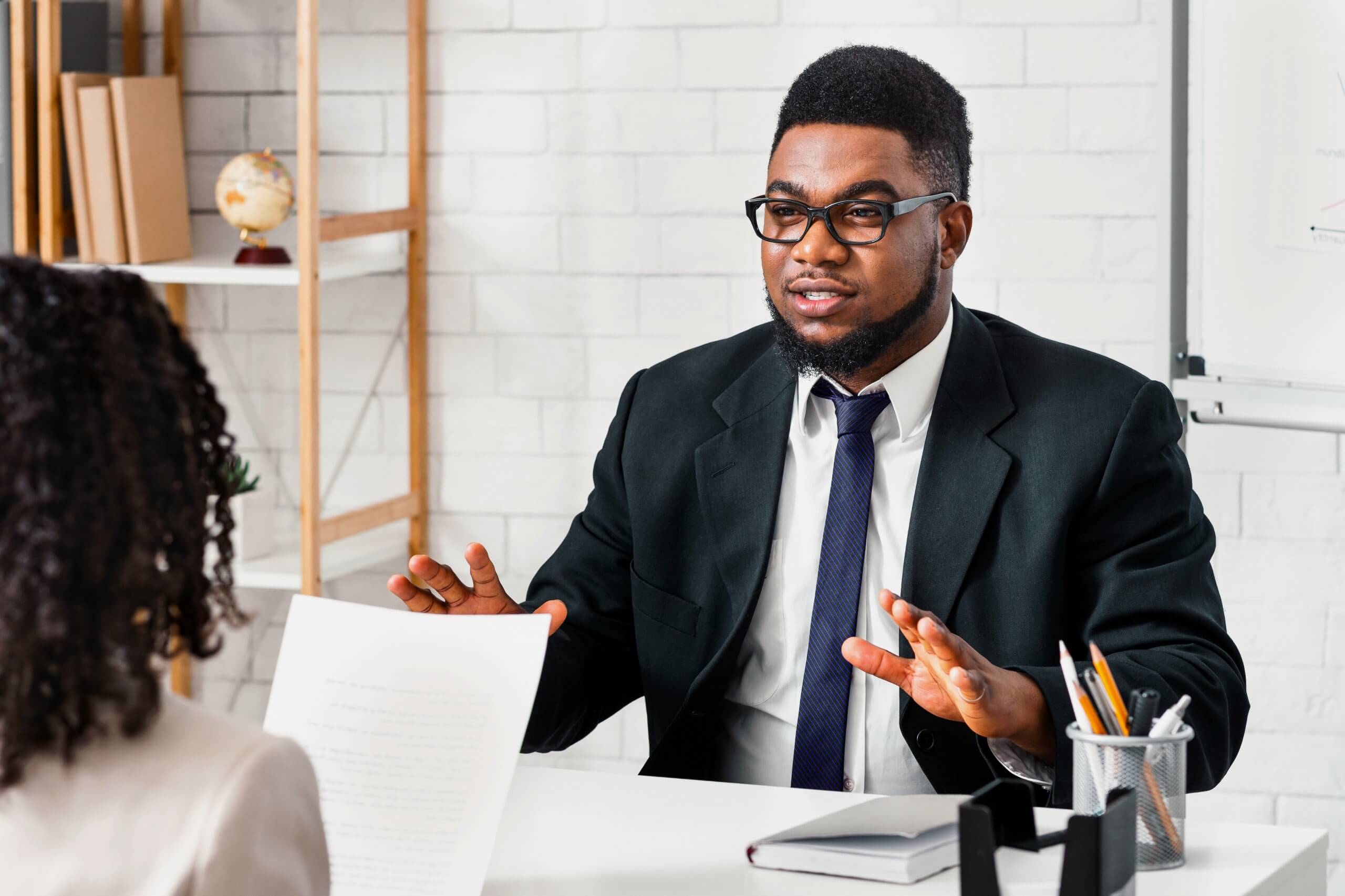A man in a suit and glasses gestures while speaking to a woman across a desk in an office, suggesting business conversations or a job interview. Papers, a notebook, and a pen holder are on the desk.