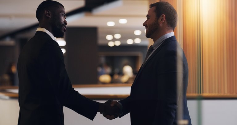 Two men in business suits shake hands in an office setting, facing each other and smiling, demonstrating leadership amid blurred lights and furniture in the background.