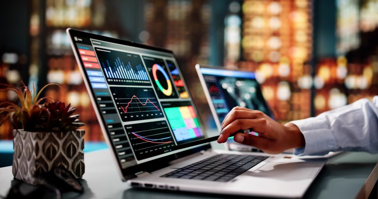 A person using a laptop displaying colorful financial charts and graphs, with another laptop, a small potted plant, and a blurred cityscape backdrop—showcasing data analysis in the Public Sector.