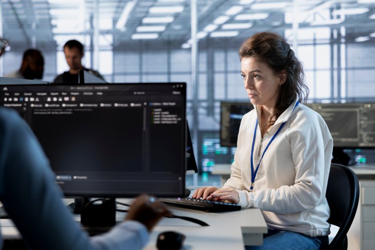A woman with a badge types on a keyboard at a desk with multiple computer monitors in a modern, well-lit public sector office while other people work in the background.