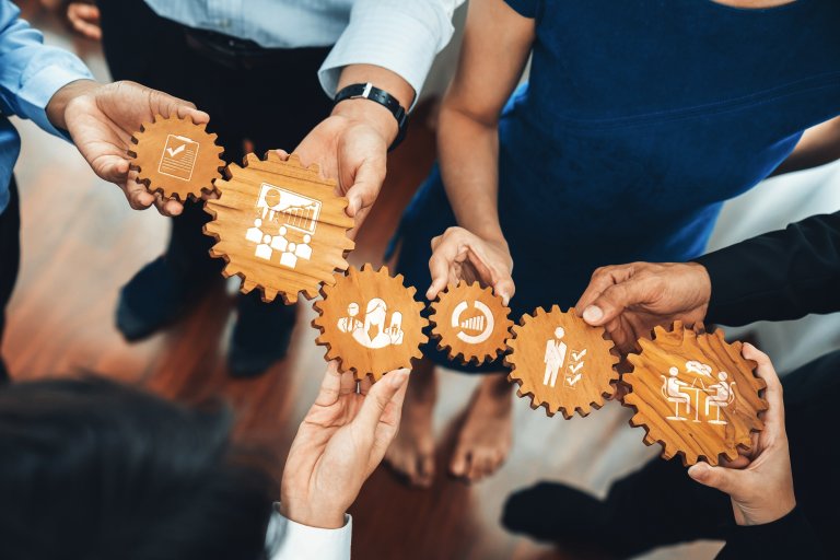 A group of people standing in a circle hold wooden gears with business-related icons, symbolizing teamwork, collaboration, a strong culture, and organizational processes.