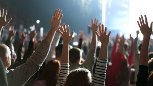 A crowd of people facing a brightly lit stage with their hands raised in the air, suggesting excitement, participation, and a shared sense of success at a concert or live event.