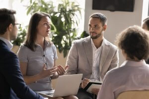 A group of four people in business attire are engaged in discussion, possibly about a Public Sector project. A woman with a laptop is speaking as others listen attentively, with green plants visible in the background.