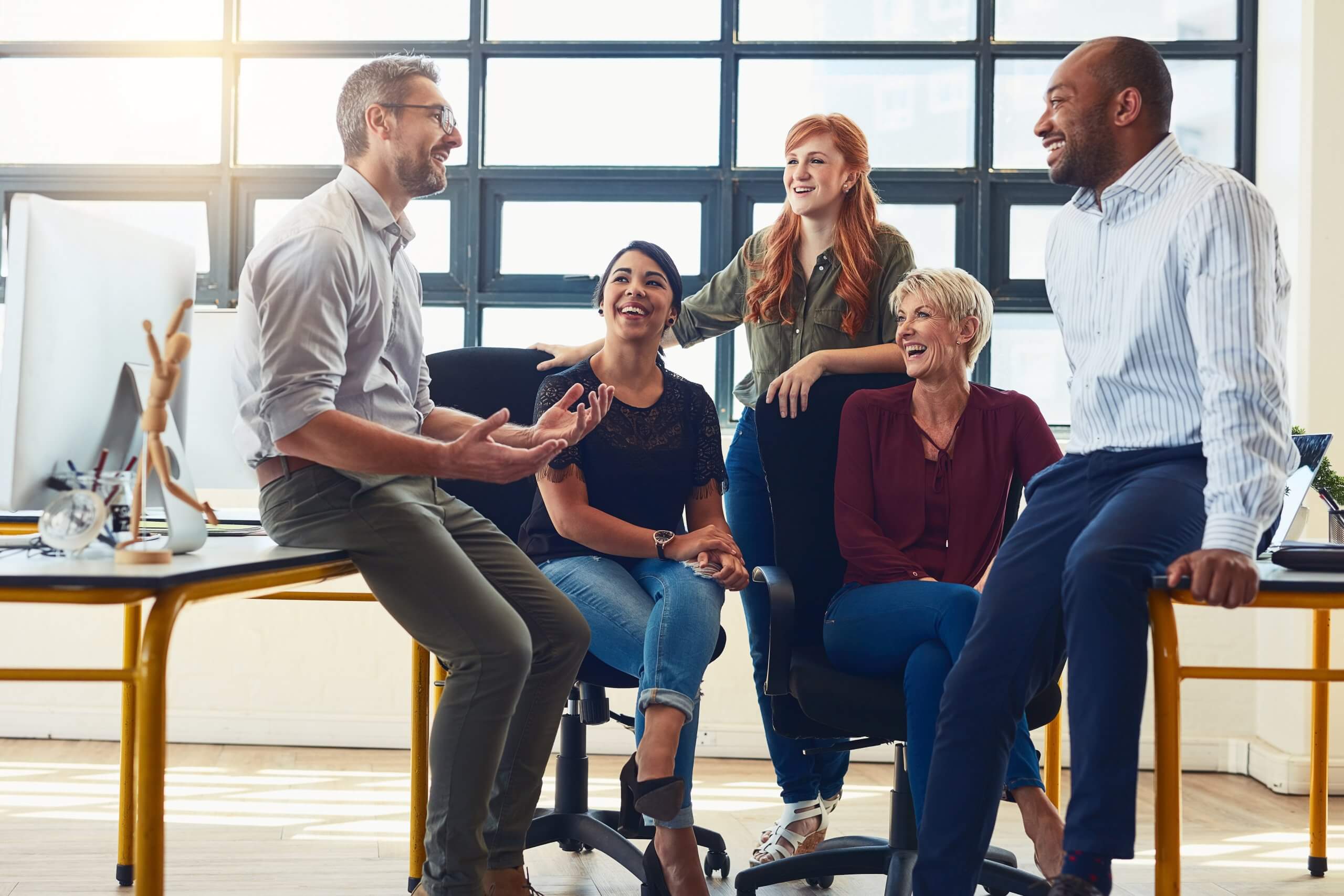 Five colleagues in casual office attire smile and laugh together in a bright office, some engaged in thinking while sitting or standing around a desk and computer, suggesting a friendly and collaborative work environment.