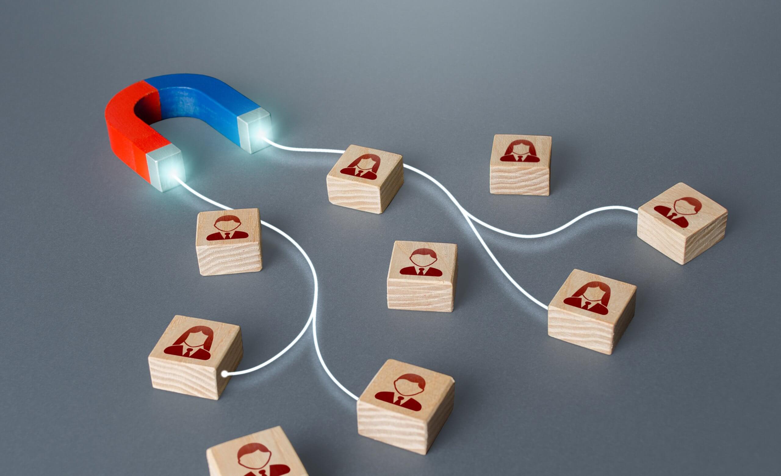 A red and blue horseshoe magnet attracts wooden blocks with male and female business icons, linked by white lines, symbolizing attracting top talent or networking.