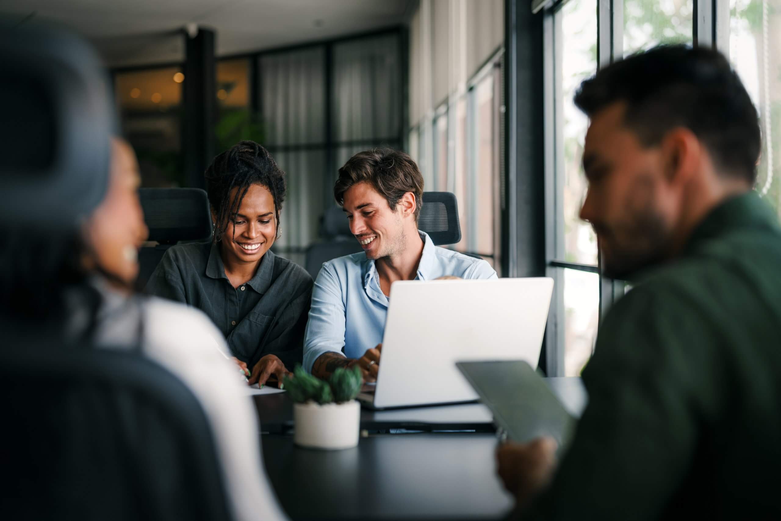 Two colleagues sit at a table with a laptop, smiling and writing notes as they engage in thinking together, while two others sit nearby, all in a modern, well-lit office with glass walls.