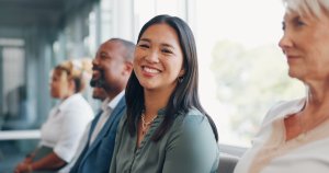 A woman smiles at the camera while sitting in a row with three other people, all dressed in business attire, during a government hiring session in a bright office setting.
