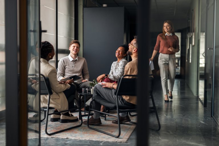 Five people in business attire are engaged in lively discussion and laughter, clearly enjoying some creative thinking in an office meeting room; one person is approaching the group, holding a notebook.