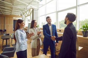 Three people stand and applaud as two talented men in business attire shake hands in a modern, sunlit office with large windows and plants, suggesting a successful business meeting or agreement.