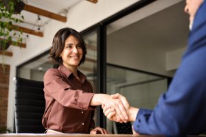 A woman in business attire smiles while shaking hands with another person across a desk in a modern office, reflecting the positive atmosphere of government hiring.