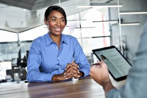 A woman in a blue shirt smiles while sitting at a table during a government hiring interview. Opposite her, a person holds a tablet displaying a digital résumé in a modern office setting.