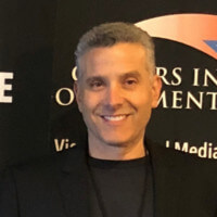 A man with short gray hair is smiling and wearing a black jacket and dark shirt, standing in front of a dark background with partially visible white and orange text about how to recruit top talent.