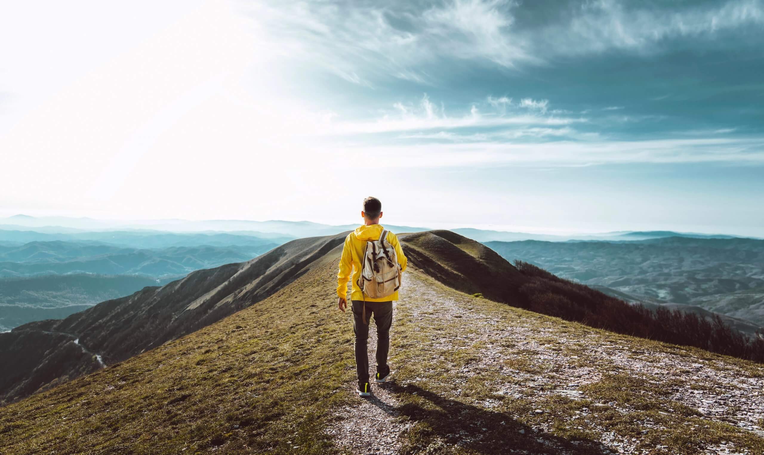 A person wearing a yellow jacket and backpack walks along a mountain ridge path, surrounded by rolling hills and valleys under a bright sky, their journey fueled by faith beneath scattered clouds.