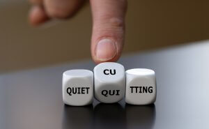 A finger points at three white dice on a table, displaying the words "QUIET QUITTING" and "CUTTING," reflecting recent trends in government hiring, with the middle die showing "CU" and "QUI.