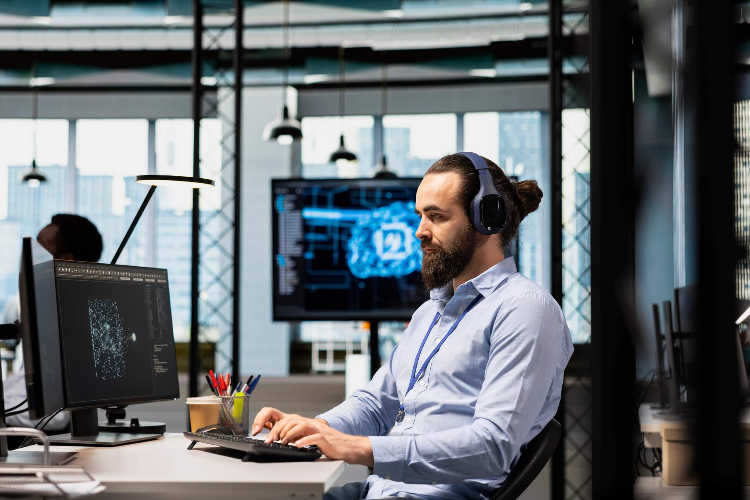 A man with a beard and headphones works on a computer in a modern office, analyzing a digital brain scan on his screen—a nod to government leadership leveraging AI innovation. A large monitor behind him displays a similar AI-themed image.