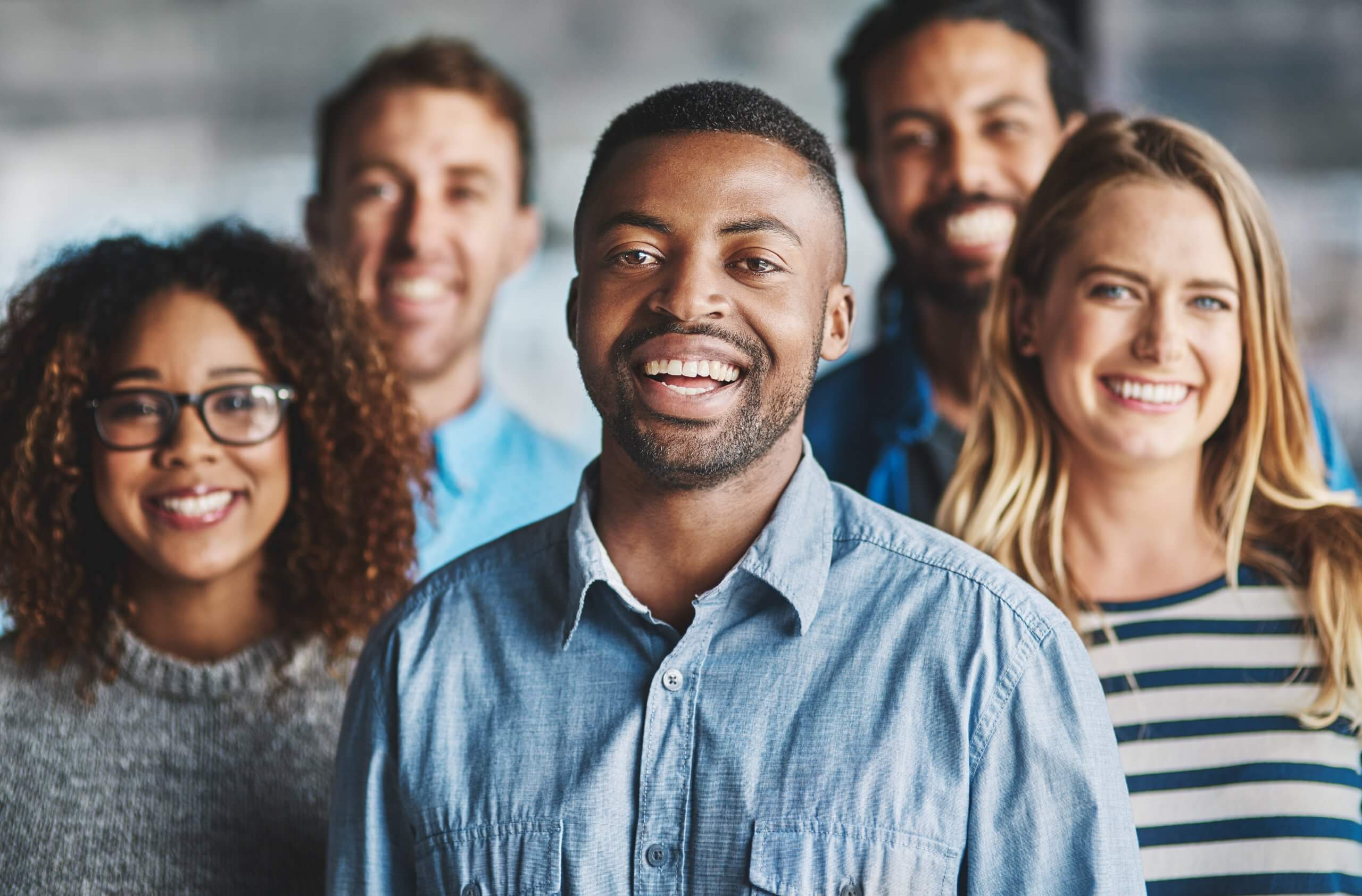 Five people stand close together, smiling at the camera. The diverse group showcases different cultures through their unique hairstyles and clothing, set against a blurred indoor background.