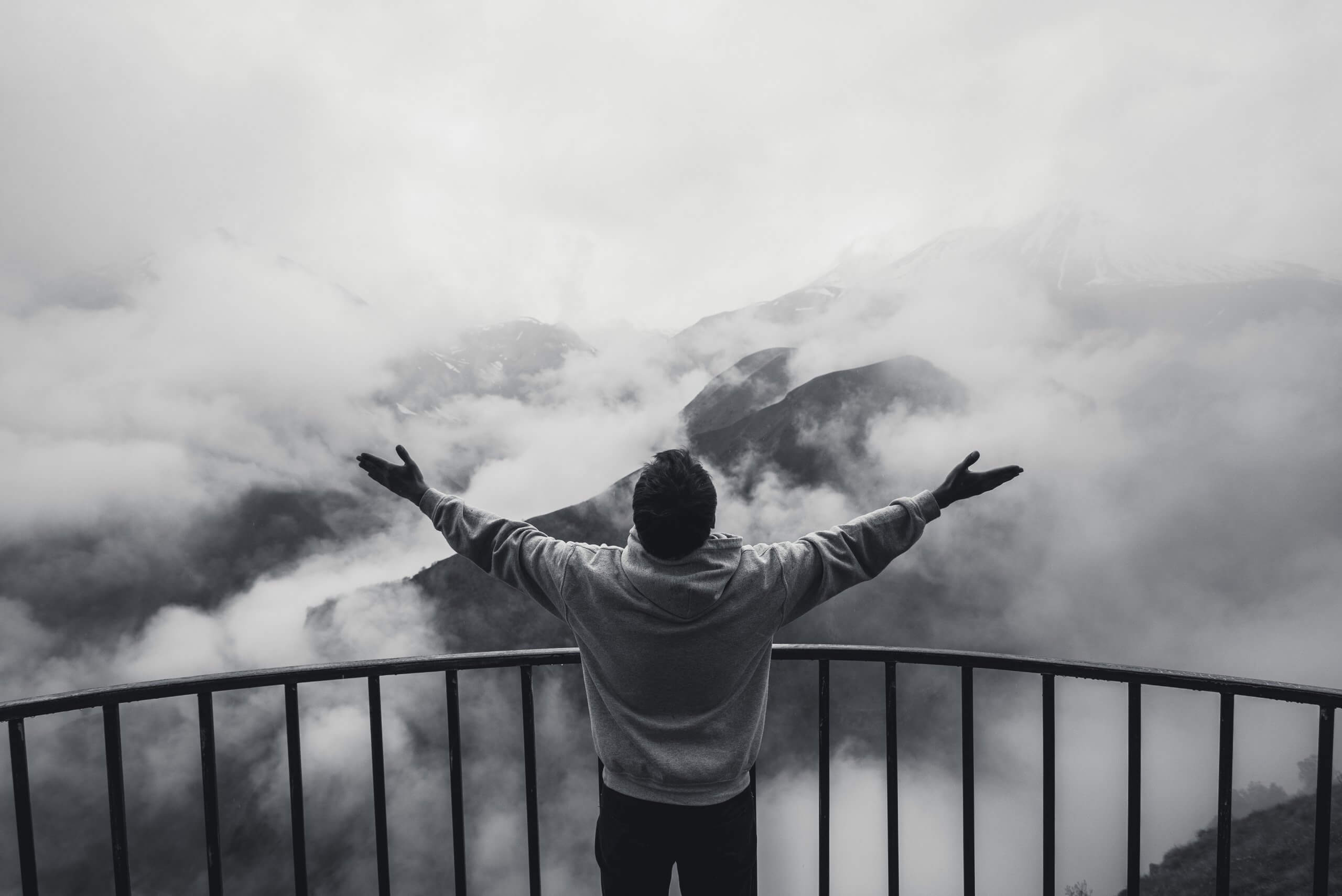 A person stands on a balcony with arms outstretched, facing foggy mountains partially covered by clouds, as if embracing the unknown with faith. The scene is in black and white, creating a dramatic and serene atmosphere.