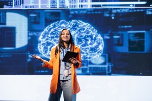 A woman in an orange blazer holds a tablet and speaks on stage, showcasing leadership as she stands before a large digital screen with a glowing brain image in the background.