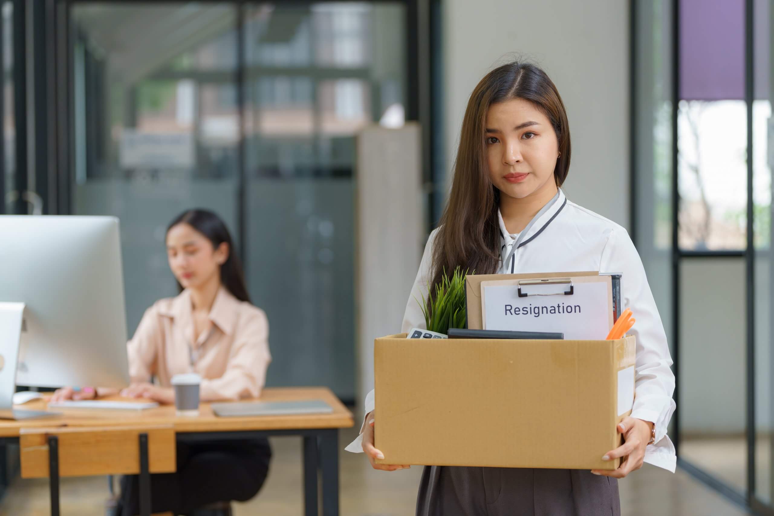 Unemployed,,Quiet,Job,,Desperate,Asian,Young,Businesswoman,Resigning,From,Company A confident man in a suit stands in the foreground of a modern office, holding glasses. In the background, two people shake hands at a table—an ideal scene for government hiring discussions—while another person observes the meeting.