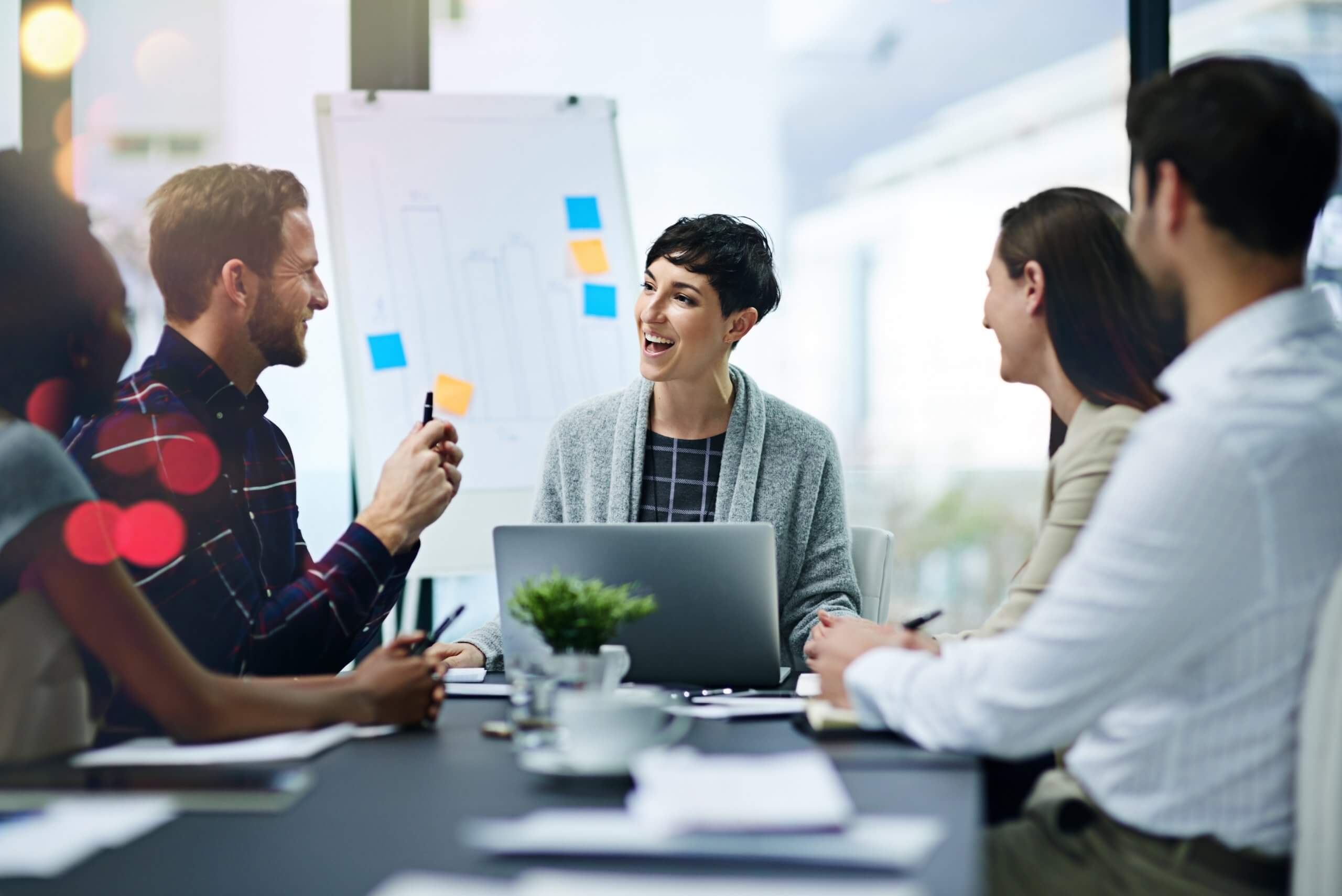 Four people sit around a table in a modern office, having a lively discussion that reflects a positive team culture. One person has a laptop open, and a flip chart with notes is in the background. They all appear engaged and smiling.