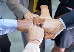 Five talented professionals stand in a circle, joining fists together in a gesture of teamwork and unity. The group is dressed in business attire, highlighting their collaborative spirit and shared talent in a professional setting.